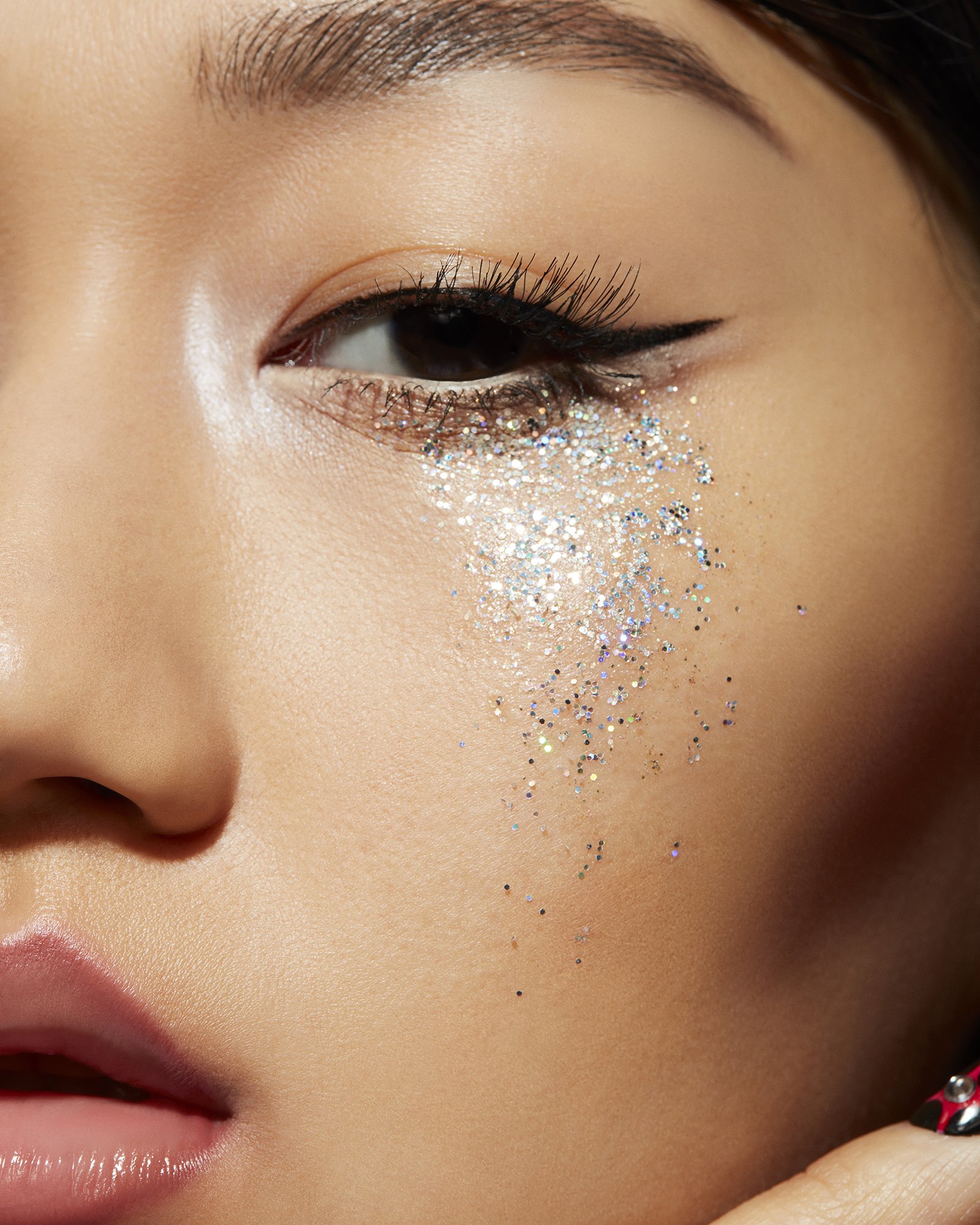 Close-up of an adult woman's face with makeup, glitter around the eye, and visible eyelashes and eyebrow in a brightly lit, indoor shot.