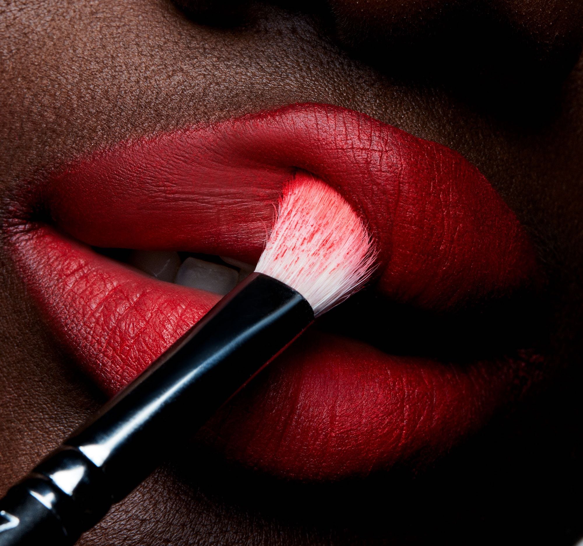 Close-up of vibrant red lipstick being applied to dark skin with a black makeup brush