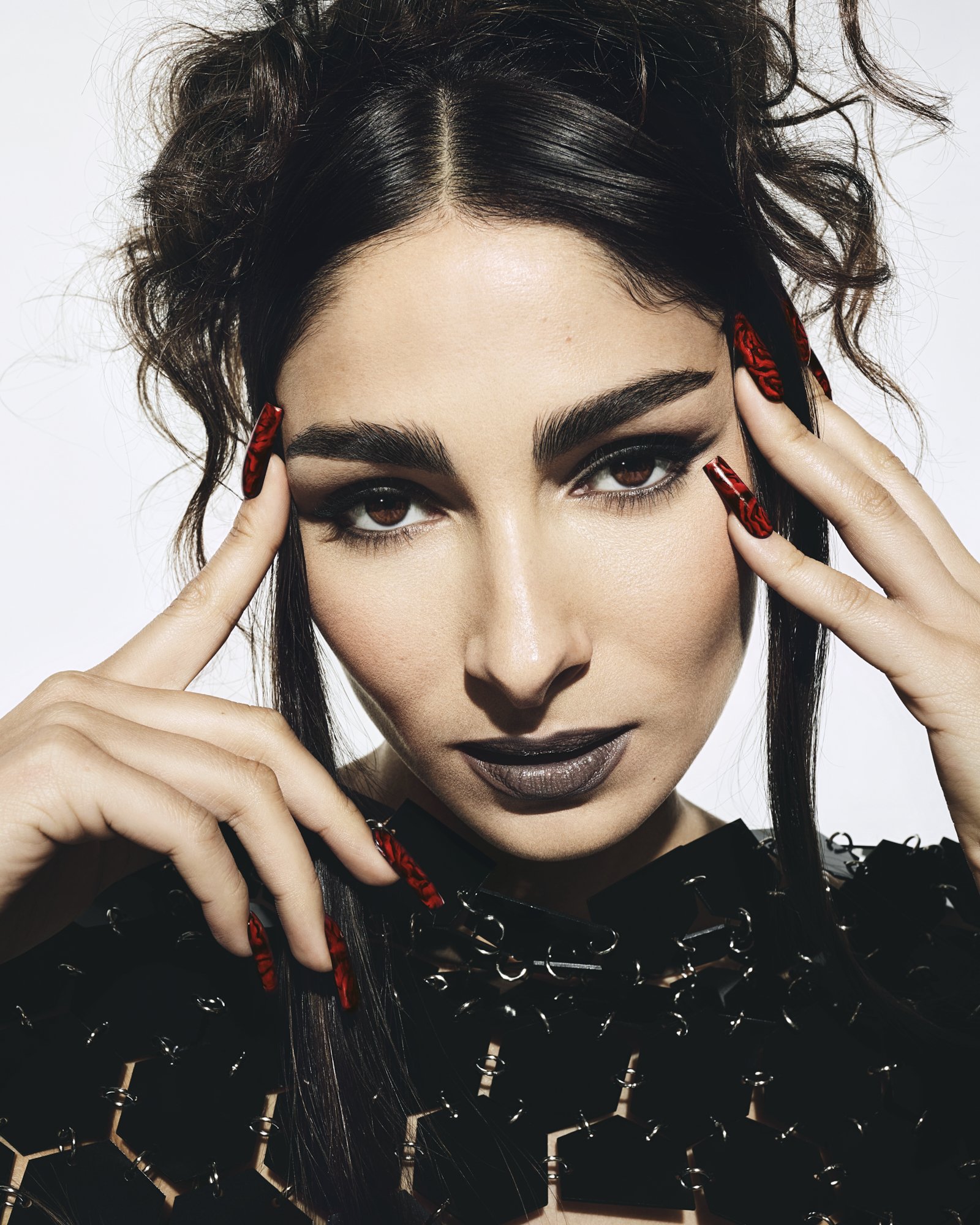 Glamorous portrait of a woman with dramatic black eye makeup and dark lipstick, holding hands near her temples against a white backdrop.