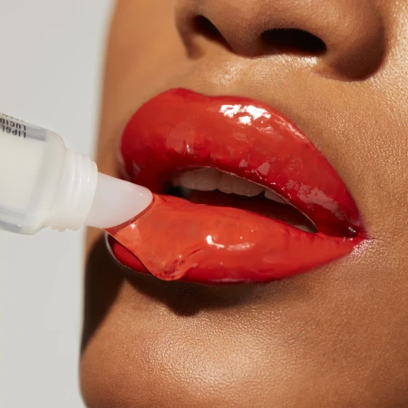 Close-up of a person applying vibrant red lip gloss with applicator to full lips. Skin is visible on face and neck, against white background.