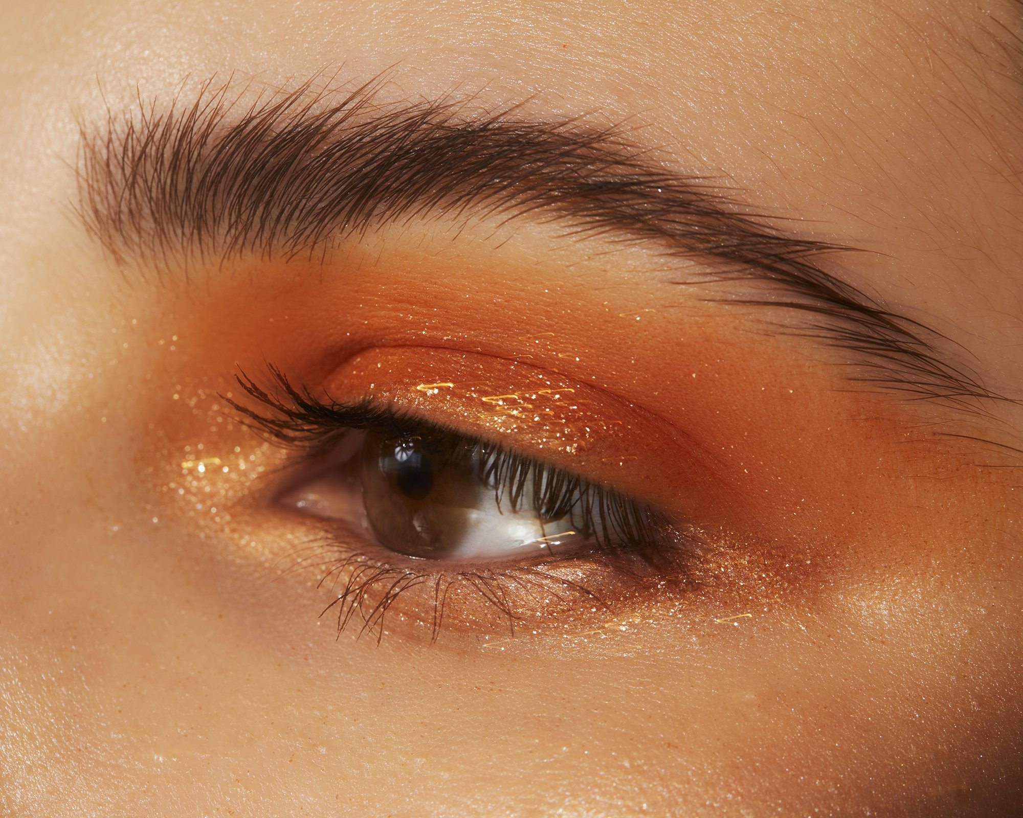 Close-up of a woman's eye with striking orange and gold shimmery eyeshadow and defined eyebrow, showcasing beauty and makeup artistry.
