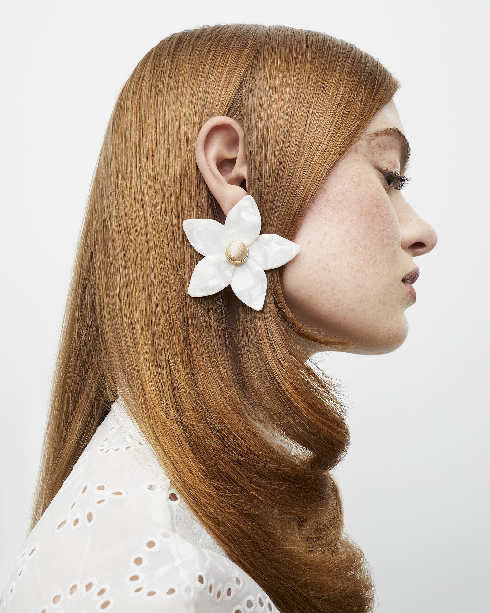Close-up of a woman with red hair wearing a large white flower earring. The earring is made of mother-of-pearl material.