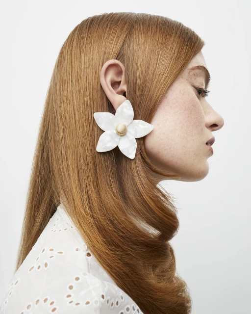 Close-up of a woman with red hair wearing a large white flower earring. The earring is made of mother-of-pearl material.