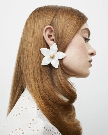 Close-up of a woman with red hair wearing a large white flower earring. The earring is made of mother-of-pearl material.