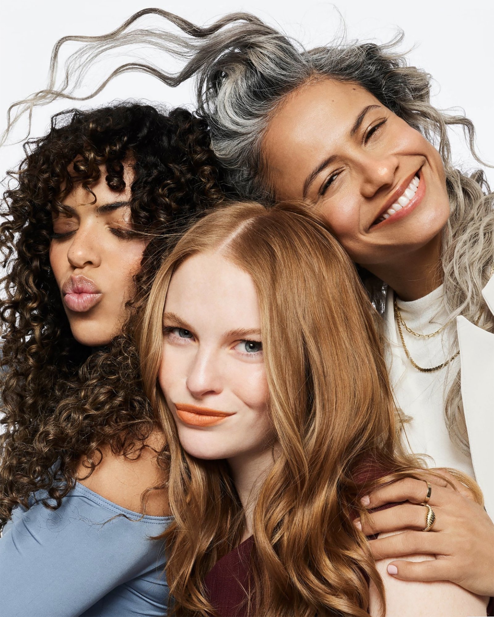 Three women of different ethnicities and hair colors pose closely together in a beauty shot against a bright white backdrop.