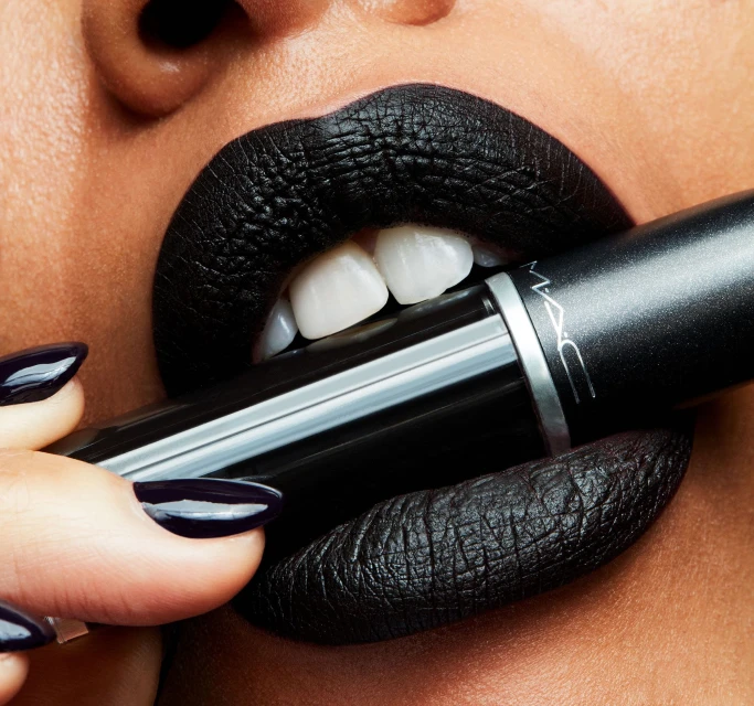 Close-up of a person's face with black lipstick and fingernails, holding a tube of MAC lipstick. Beauty shot for make-up and cosmetic advertising.
