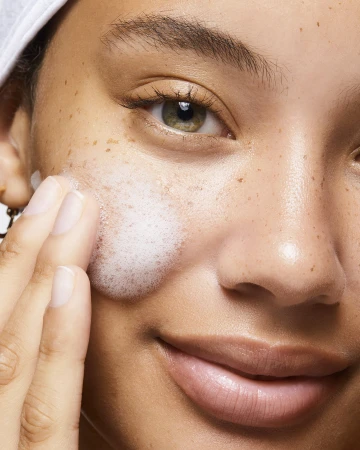 Close-up of a woman with fair skin and freckles gently applying white, foamy cleanser to her face, showcasing skincare routine.