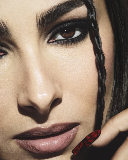 Close-up of a woman's face with dramatic black eye makeup, a single braid framing her face, and a fingernail with a rose design.