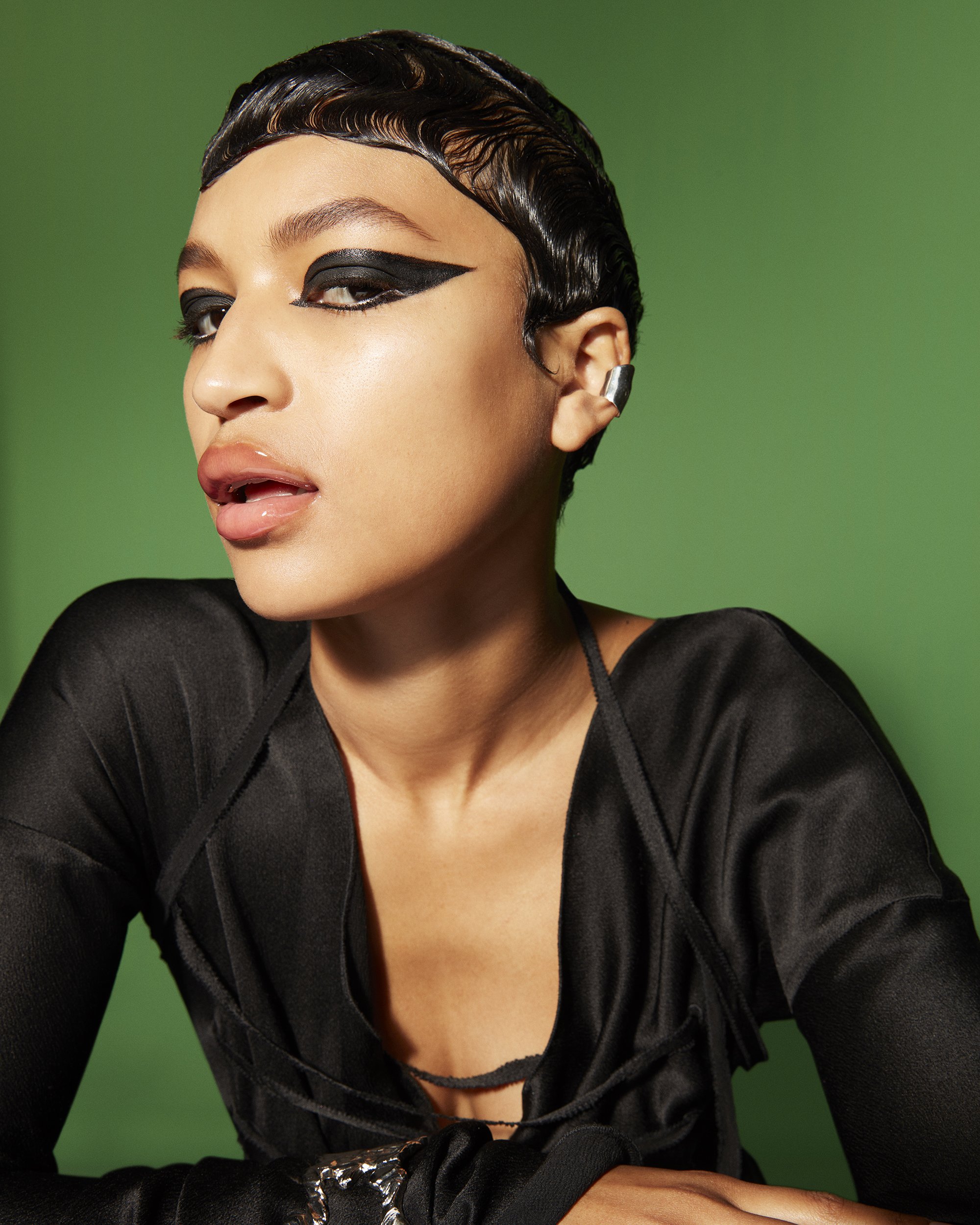 Portrait of an African-descent adult woman with dark waved hair, a black outfit, silver ear cuff, and dramatic winged black eye makeup.