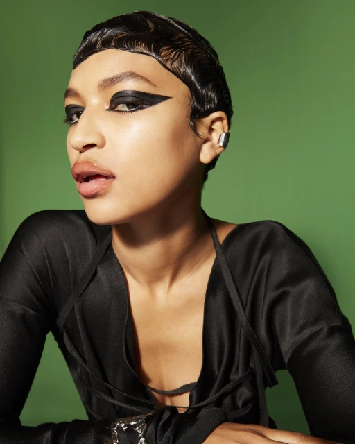Portrait of an African-descent adult woman with dark waved hair, a black outfit, silver ear cuff, and dramatic winged black eye makeup.