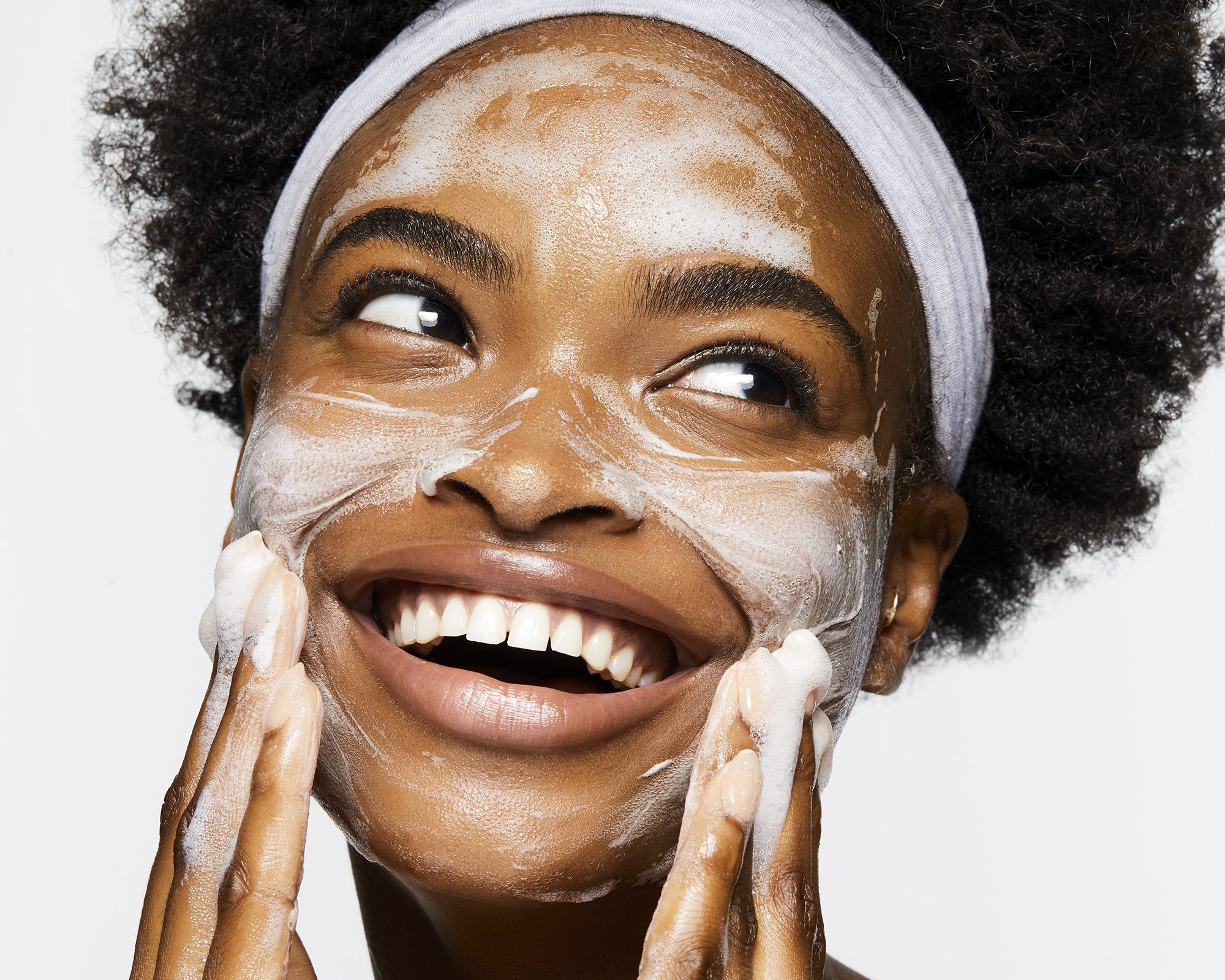 Close-up studio shot of an African American woman with natural hair smiling while applying facial cleanser. Beauty and skincare concept.