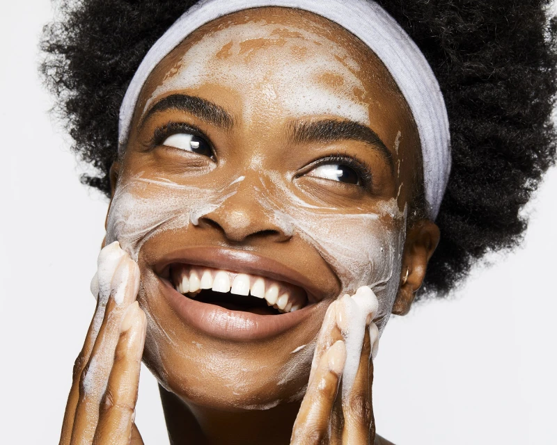 Close-up studio shot of an African American woman with natural hair smiling while applying facial cleanser. Beauty and skincare concept.