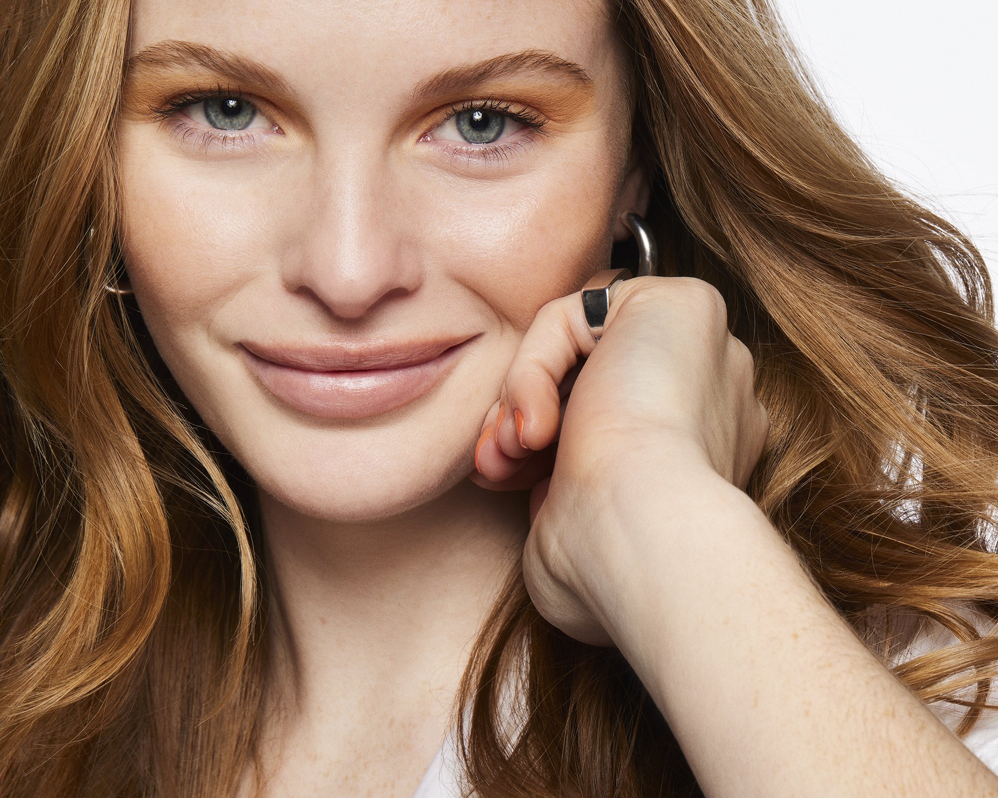 Close-up portrait of a fair-skinned woman with light red hair smiling slightly, wearing silver hoop earrings, against a white backdrop.