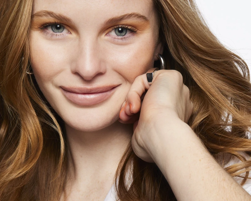 Close-up portrait of a fair-skinned woman with light red hair smiling slightly, wearing silver hoop earrings, against a white backdrop.