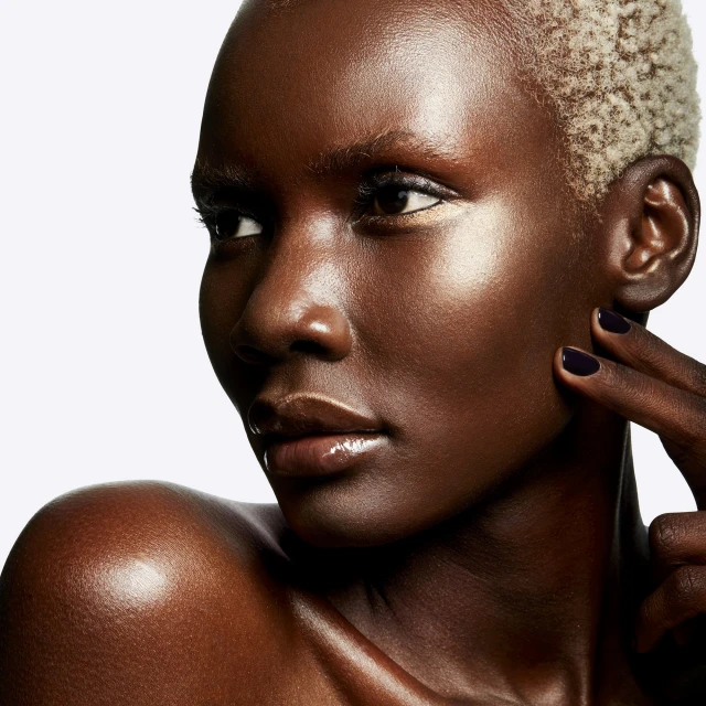 Close-up of an adult female with short blonde hair and dark skin, her radiant face and glossy lips catching the light on a white background.