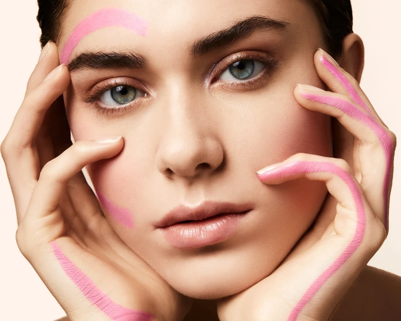 Close-up portrait of a woman with pink makeup on her face and hands, set against a soft cream backdrop in a beauty context.