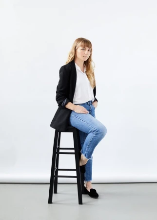 Woman in black blazer and blue jeans seated on a black stool against a white seamless background