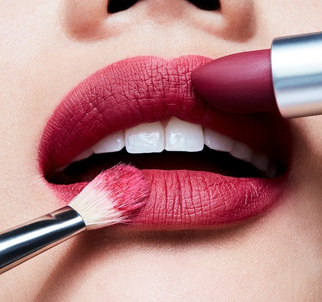 Close-up shot of a person's lips with red lipstick being applied using a lipstick tube and a makeup brush, beauty product.