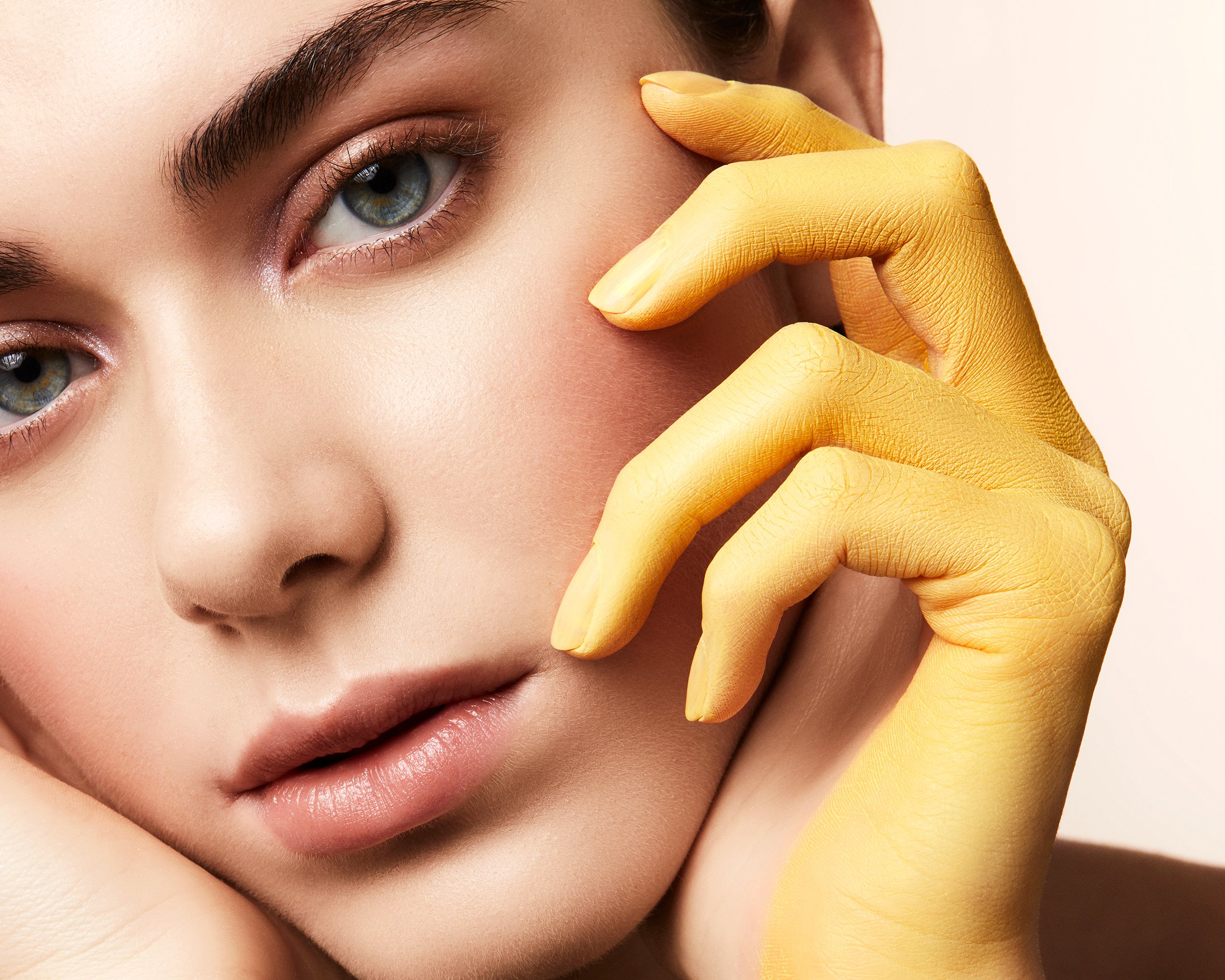 Close-up studio shot of a fair-skinned adult female model with hand painted yellow, light blue eyes, soft pink lips, and neutral expression.