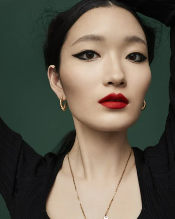 Close-up portrait of an East Asian woman with bold winged eyeliner, vibrant red lipstick, gold hoop earrings, and a gold chain necklace.