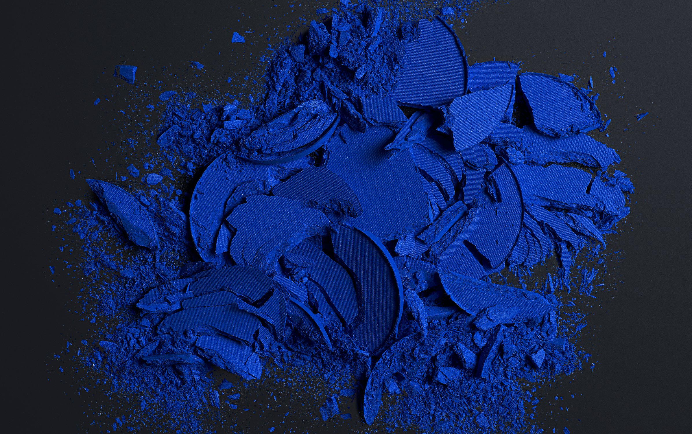 Crushed bright blue eye shadow scattered across a dark surface in a studio, creating a textured, abstract cosmetic still life.