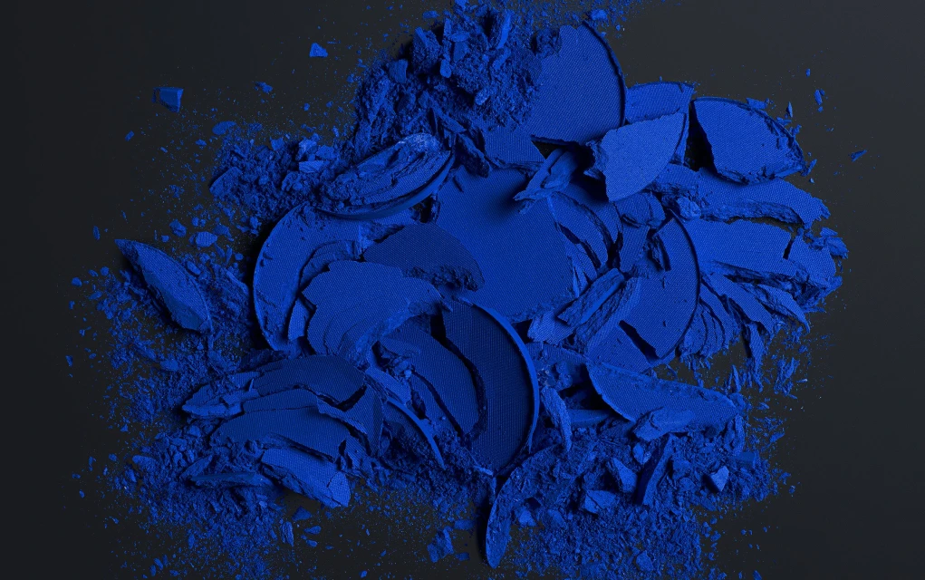 Crushed bright blue eye shadow scattered across a dark surface in a studio, creating a textured, abstract cosmetic still life.