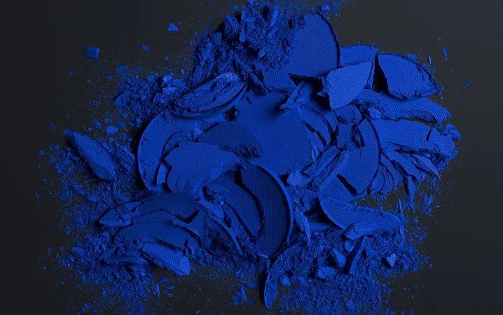 Crushed bright blue eye shadow scattered across a dark surface in a studio, creating a textured, abstract cosmetic still life.