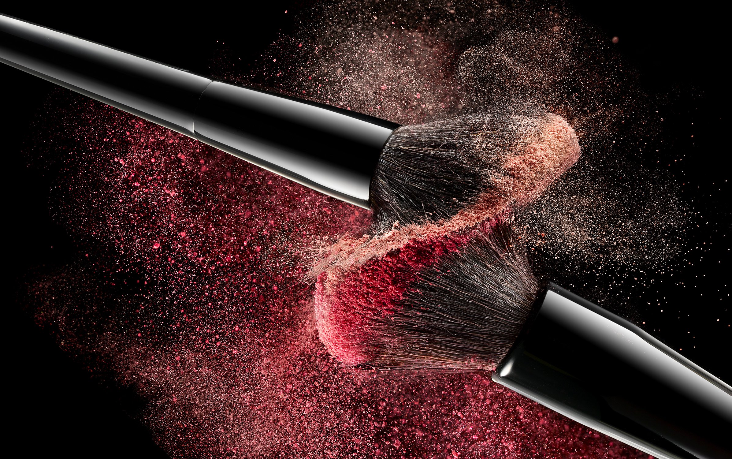 Close-up of two black makeup brushes colliding, with a burst of pink and tan powder against a dark black background.