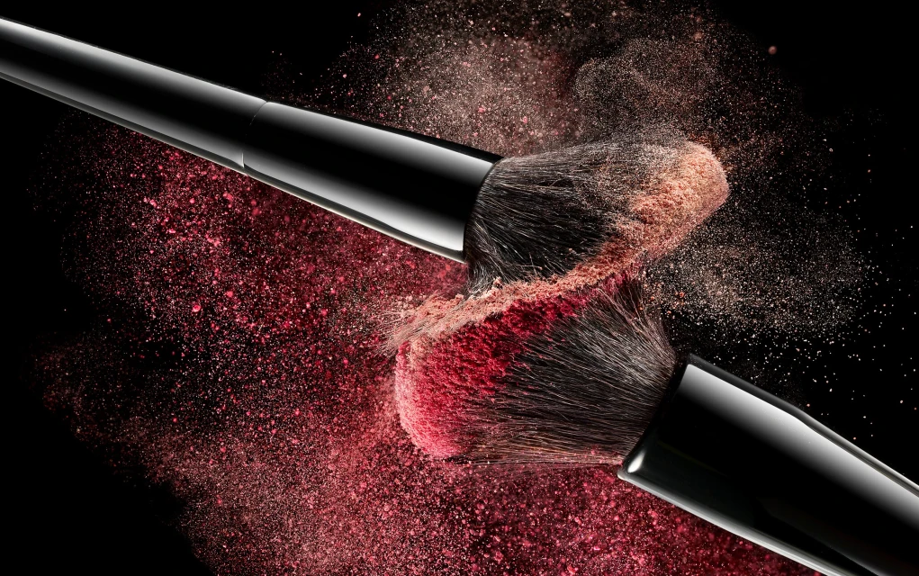 Close-up of two black makeup brushes colliding, with a burst of pink and tan powder against a dark black background.