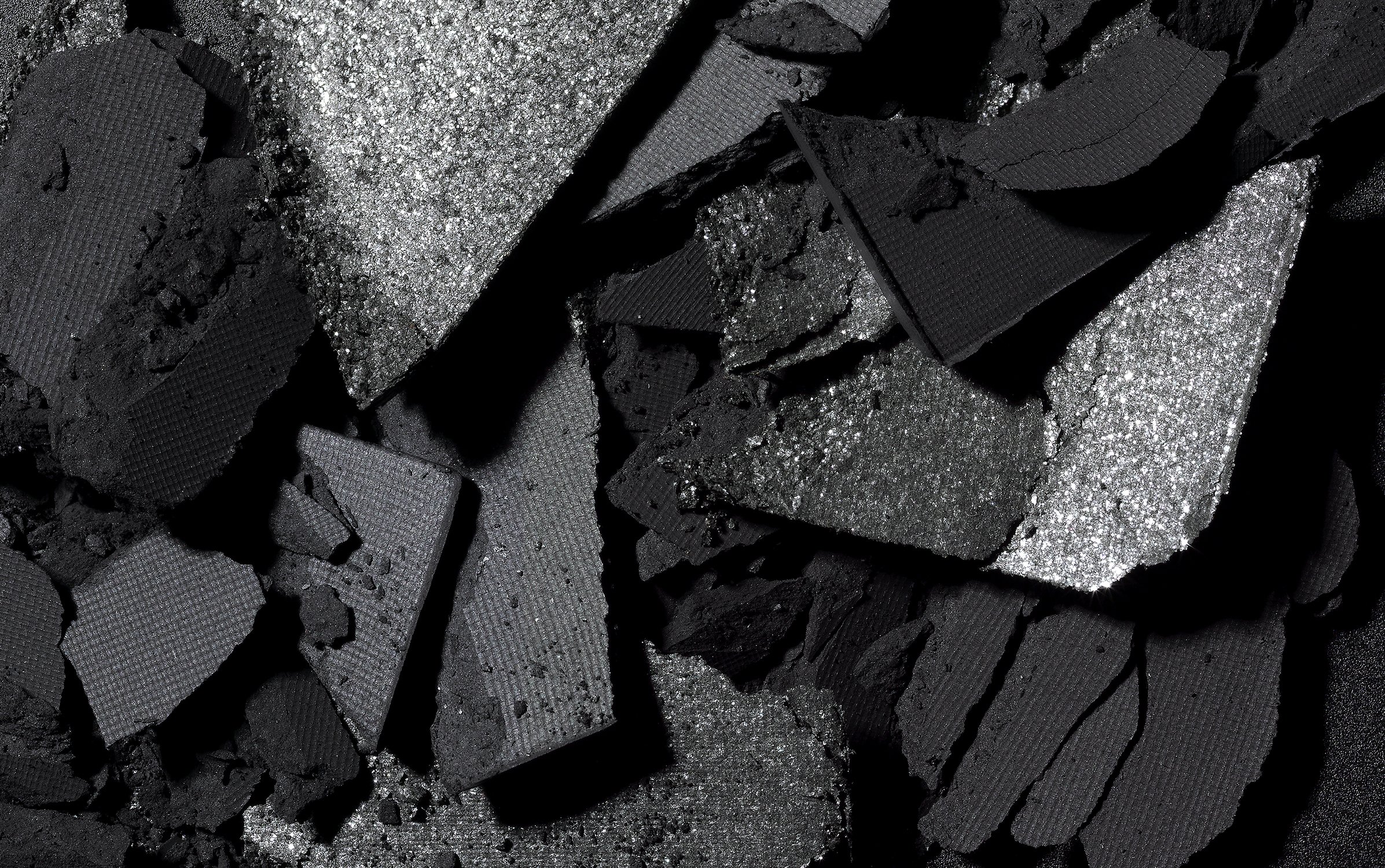 Close-up of crushed black and silver glitter eyeshadow palettes. The texture is broken and reflects light, creating a dramatic makeup look.