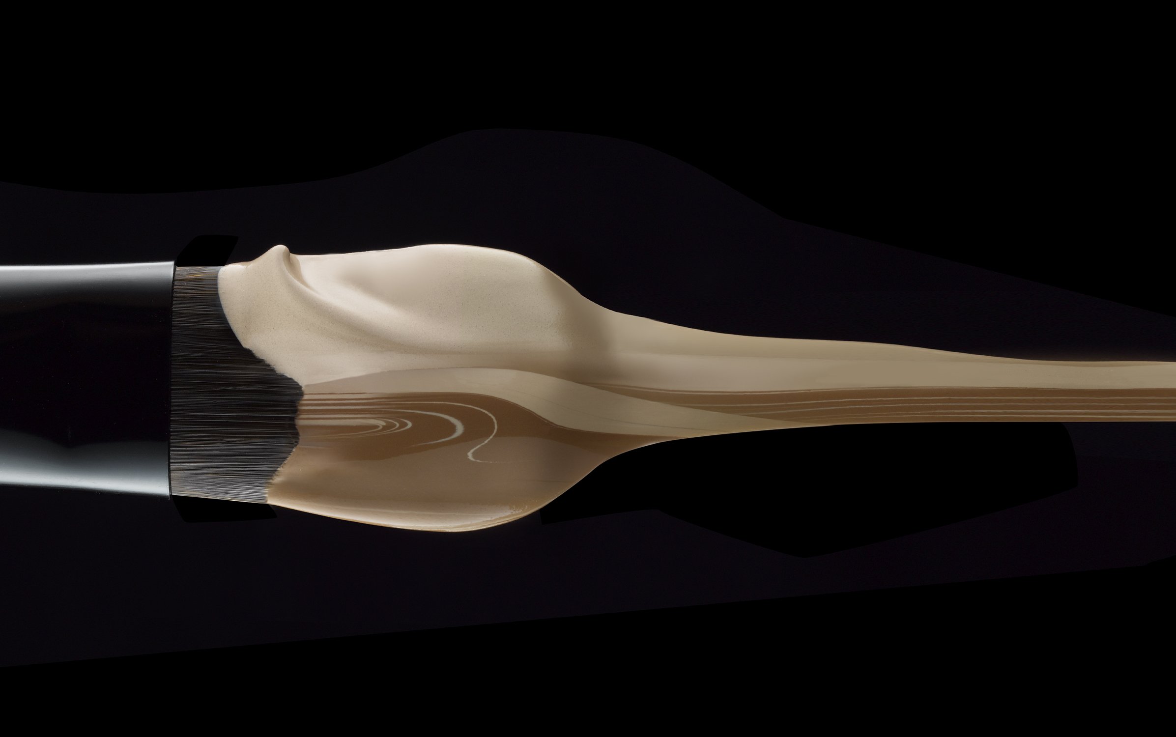 Macro photograph of foundation makeup swatches in shades of tan on a makeup brush against a stark black background.