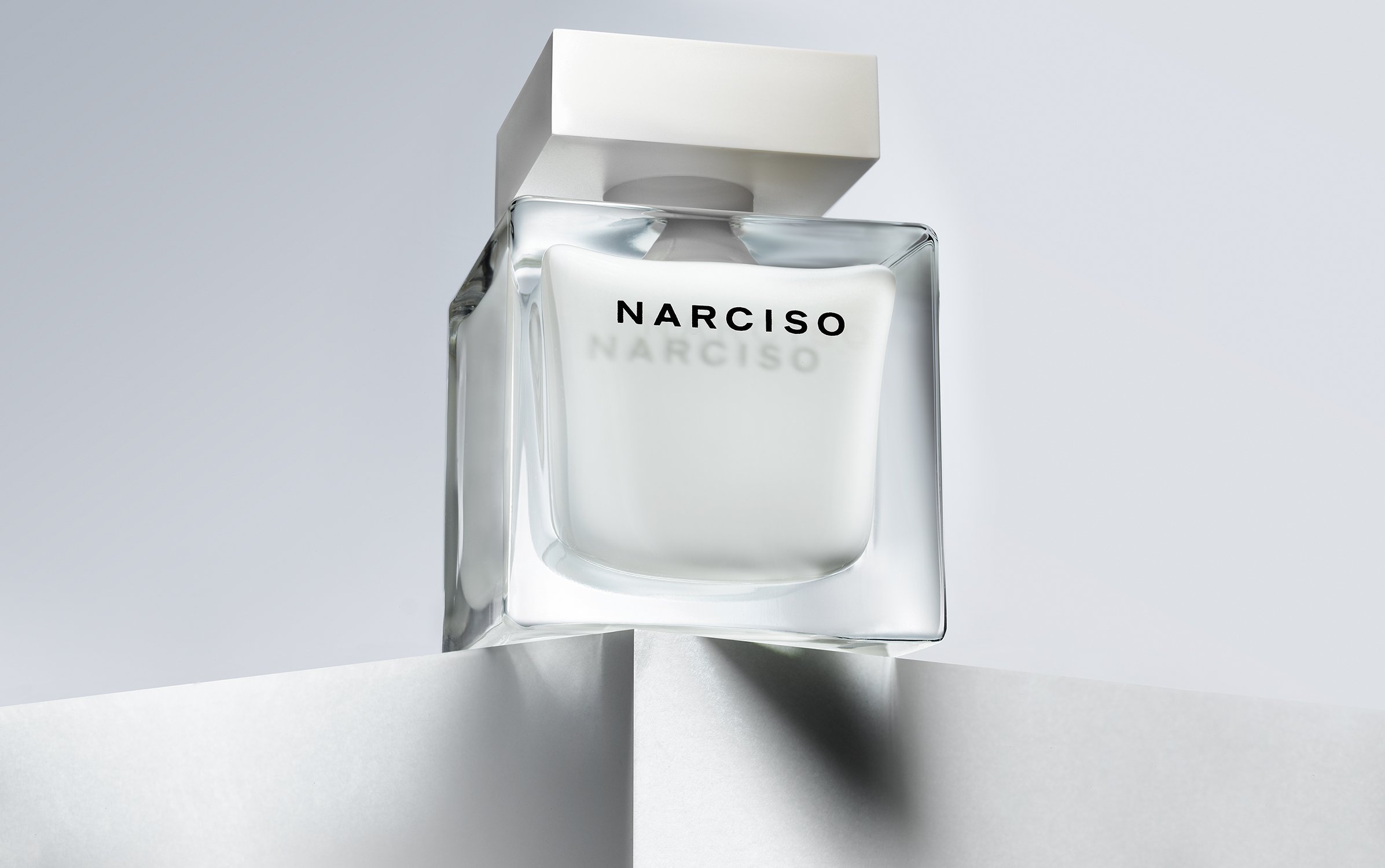 Still shot of a clear glass Narciso perfume bottle with a white cap, resting on a white geometric surface against a soft grey backdrop.