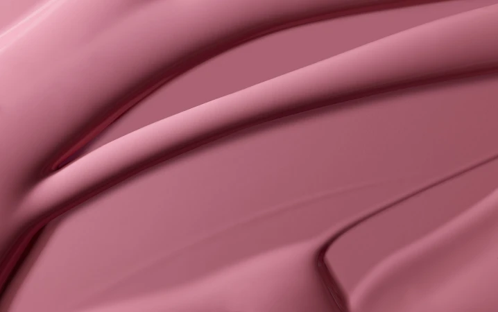 Abstract close-up shot of textured, glossy makeup in a solid color of rose pink, with swirling, rounded linear patterns and shadows.
