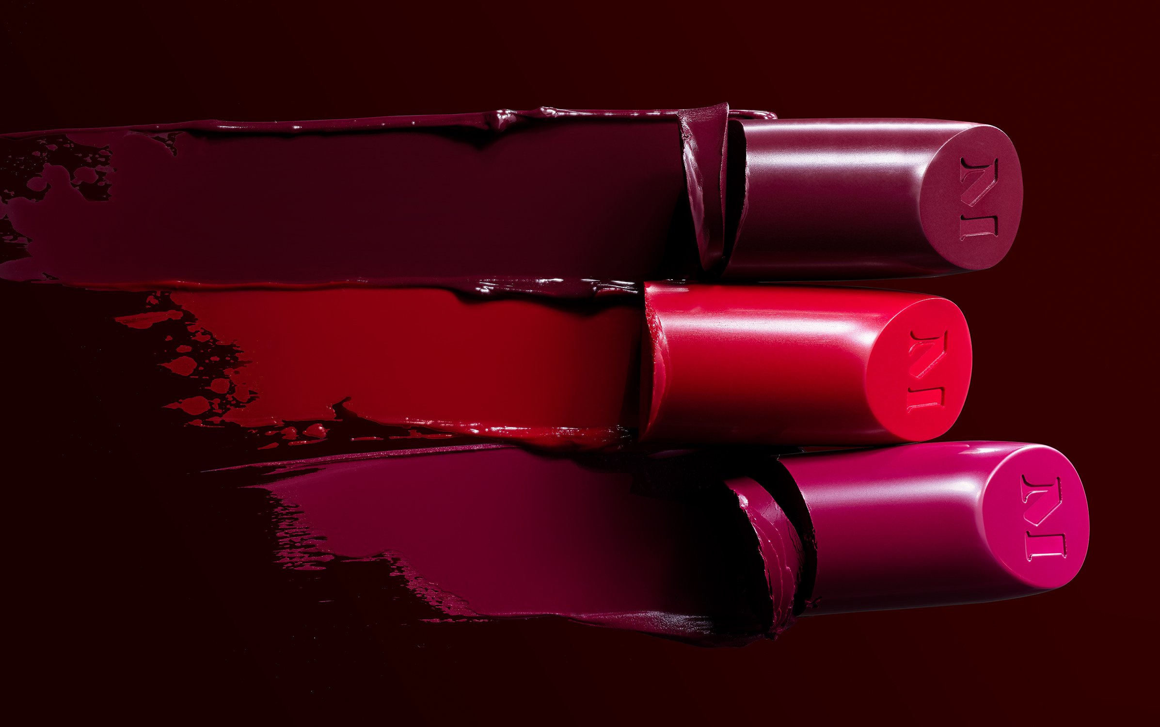 Three shades of lipstick, burgundy, red, and fuchsia, sit atop matching swipes of color, displayed against a rich maroon background.