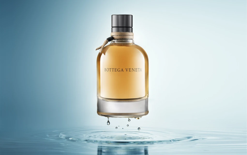 Bottega Veneta perfume bottle with amber liquid against a gradient blue backdrop and water reflections, beauty and product shot.