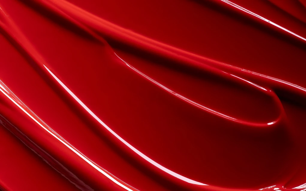 High angle view of red lipstick texture featuring a glossy, shiny surface with curved patterns of light and dark red tones.