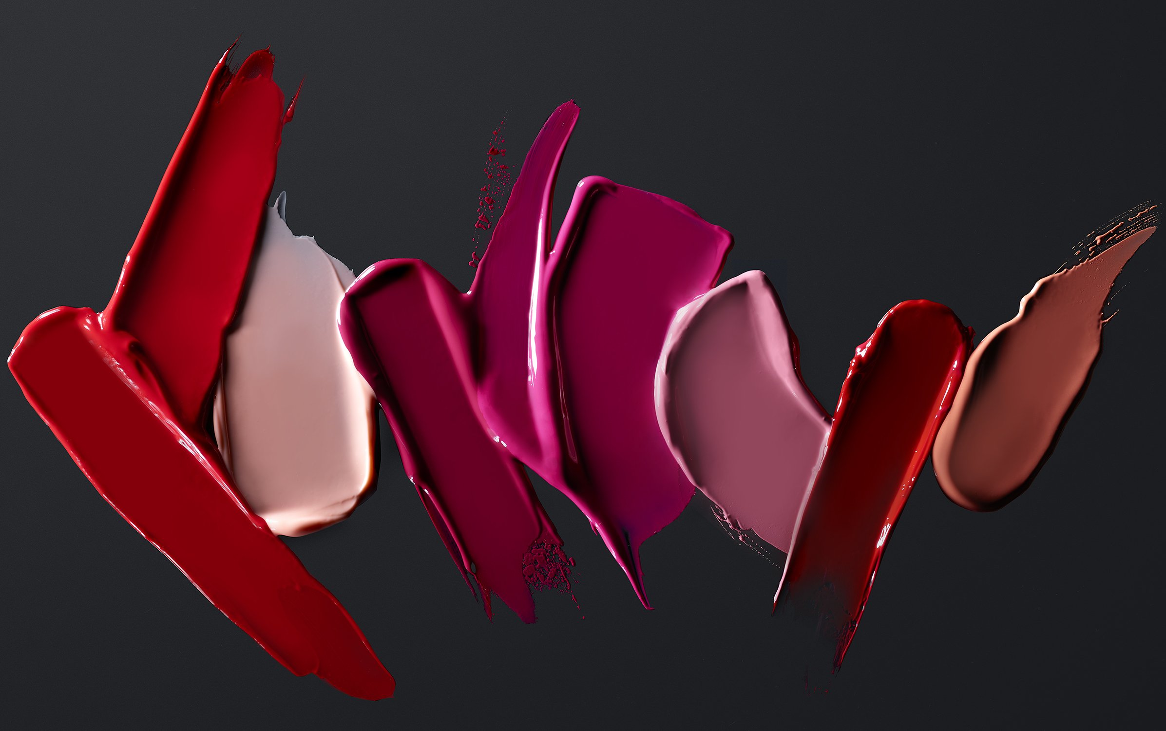 Close-up of lipstick swatches in various shades of red, pink, and beige against a dark background, showcasing beauty product texture.