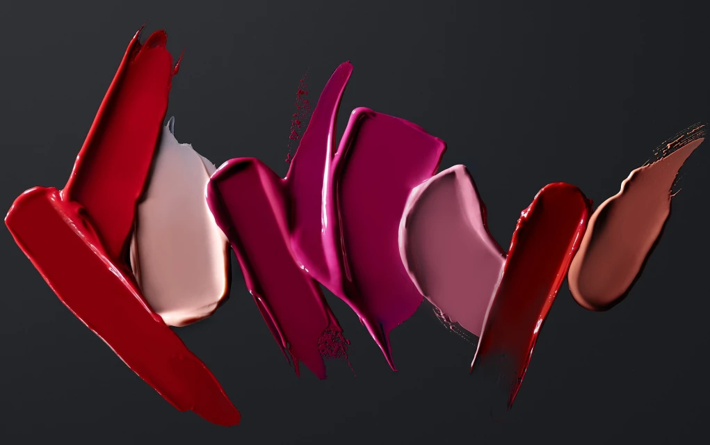 Close-up of lipstick swatches in various shades of red, pink, and beige against a dark background, showcasing beauty product texture.