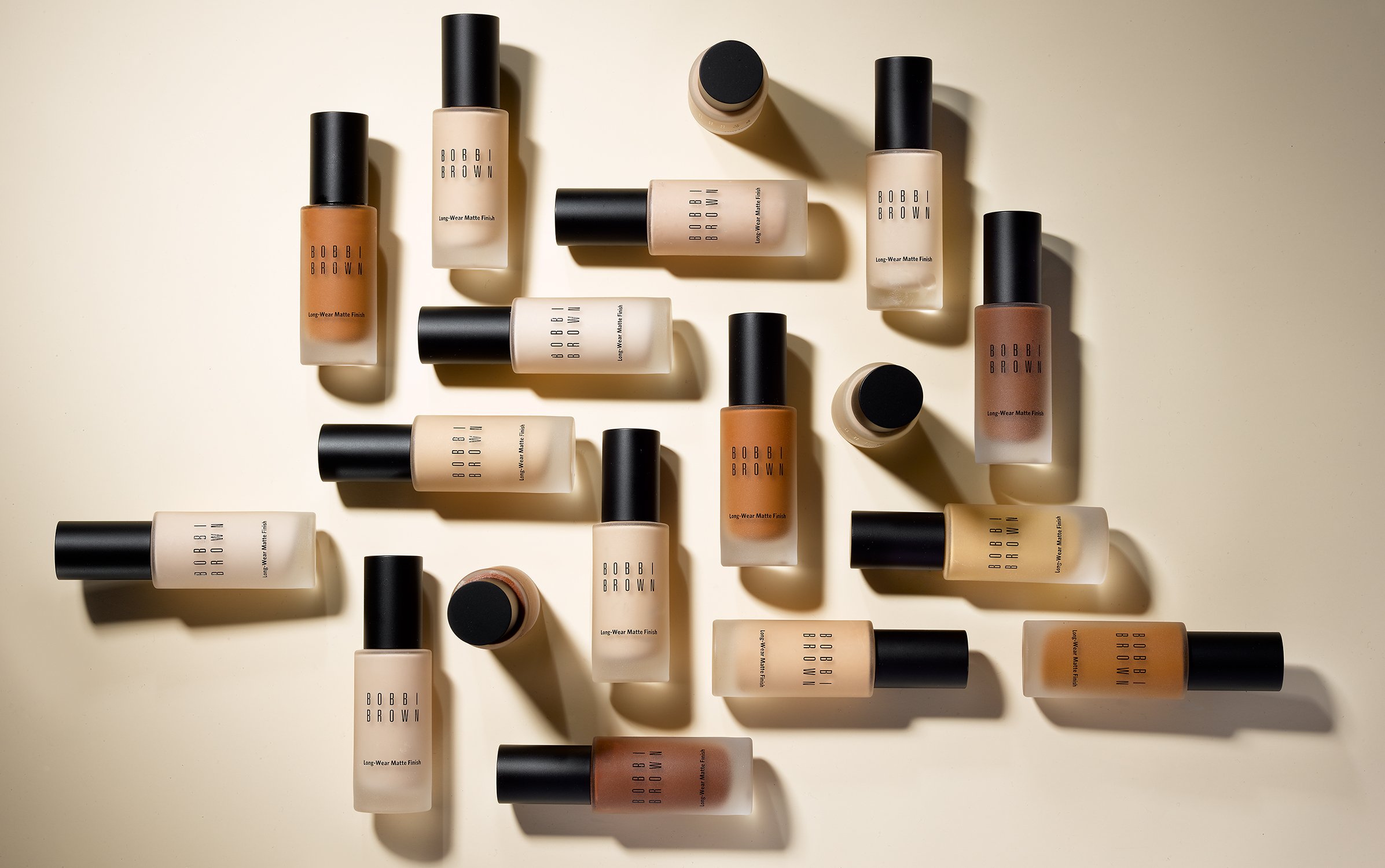 A collection of Bobbi Brown Long-Wear Matte Foundation bottles in various shades are arranged on a light background in a beauty product still life.