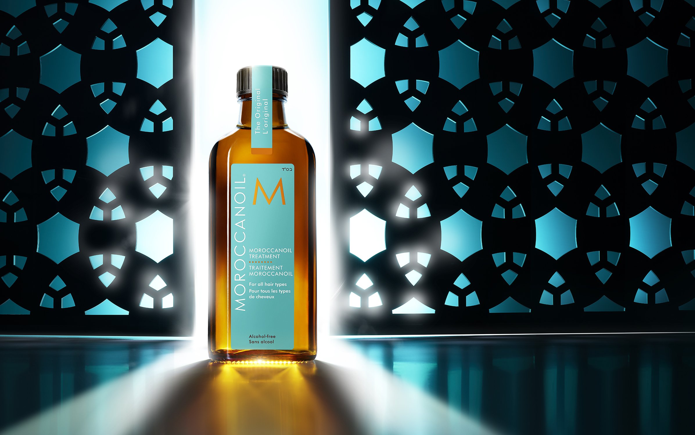 Moroccanoil treatment product shot against a geometric patterned background with teal highlights and an amber bottle.