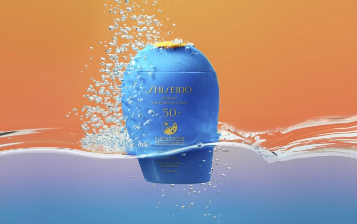 Blue bottle of Shiseido Ultimate Sun Protector Lotion SPF 50+ partially submerged in water, surrounded by bubbles against orange background.