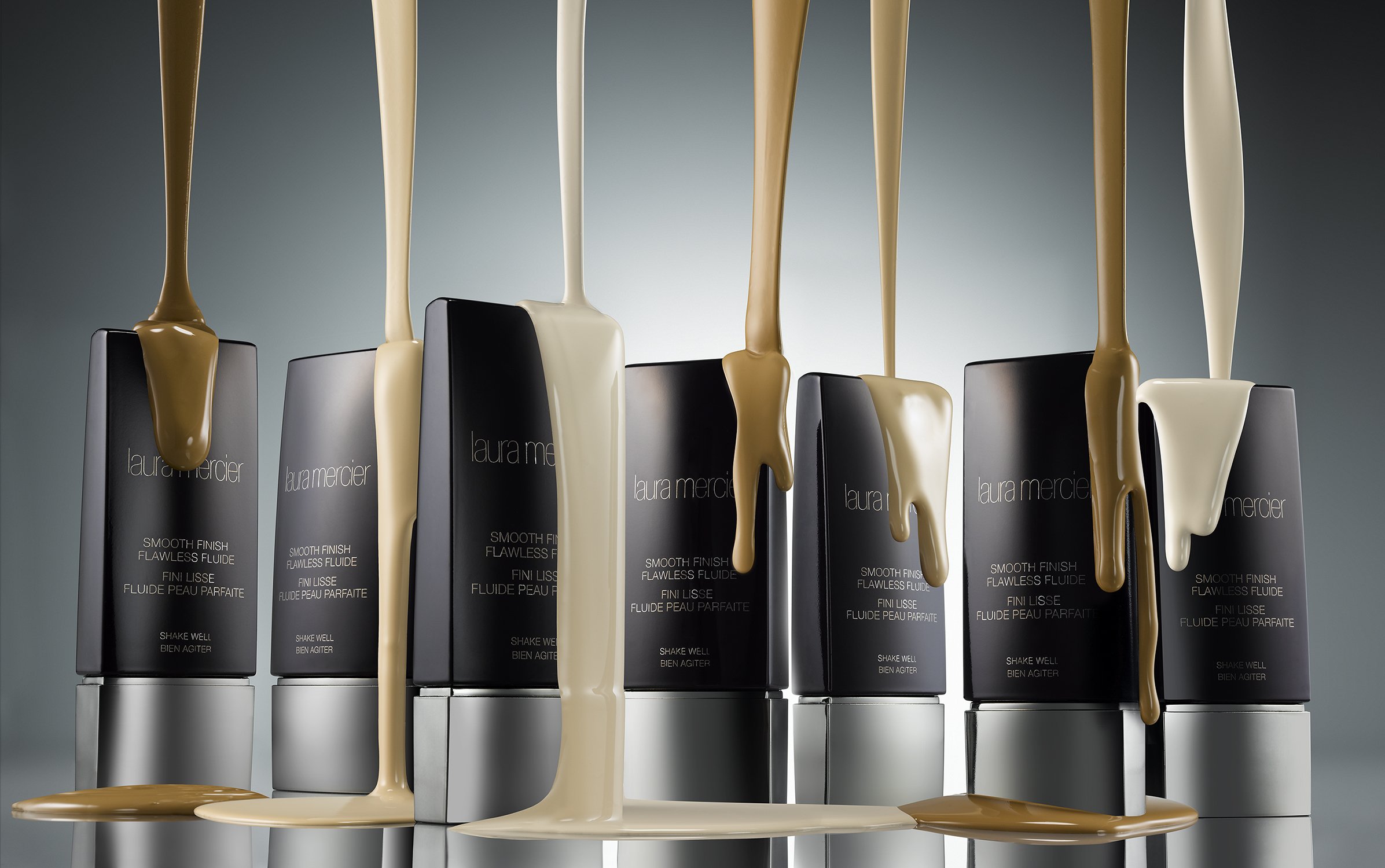 Laura Mercier Smooth Finish Flawless Fluide foundation product shot with streams of tan, beige, and white liquid makeup on containers.