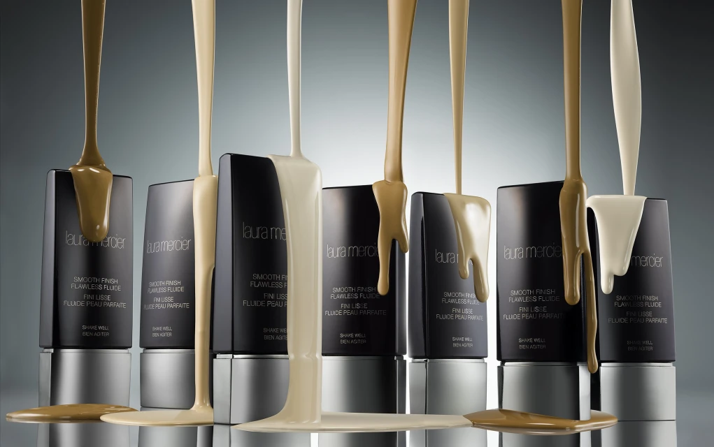 Laura Mercier Smooth Finish Flawless Fluide foundation product shot with streams of tan, beige, and white liquid makeup on containers.