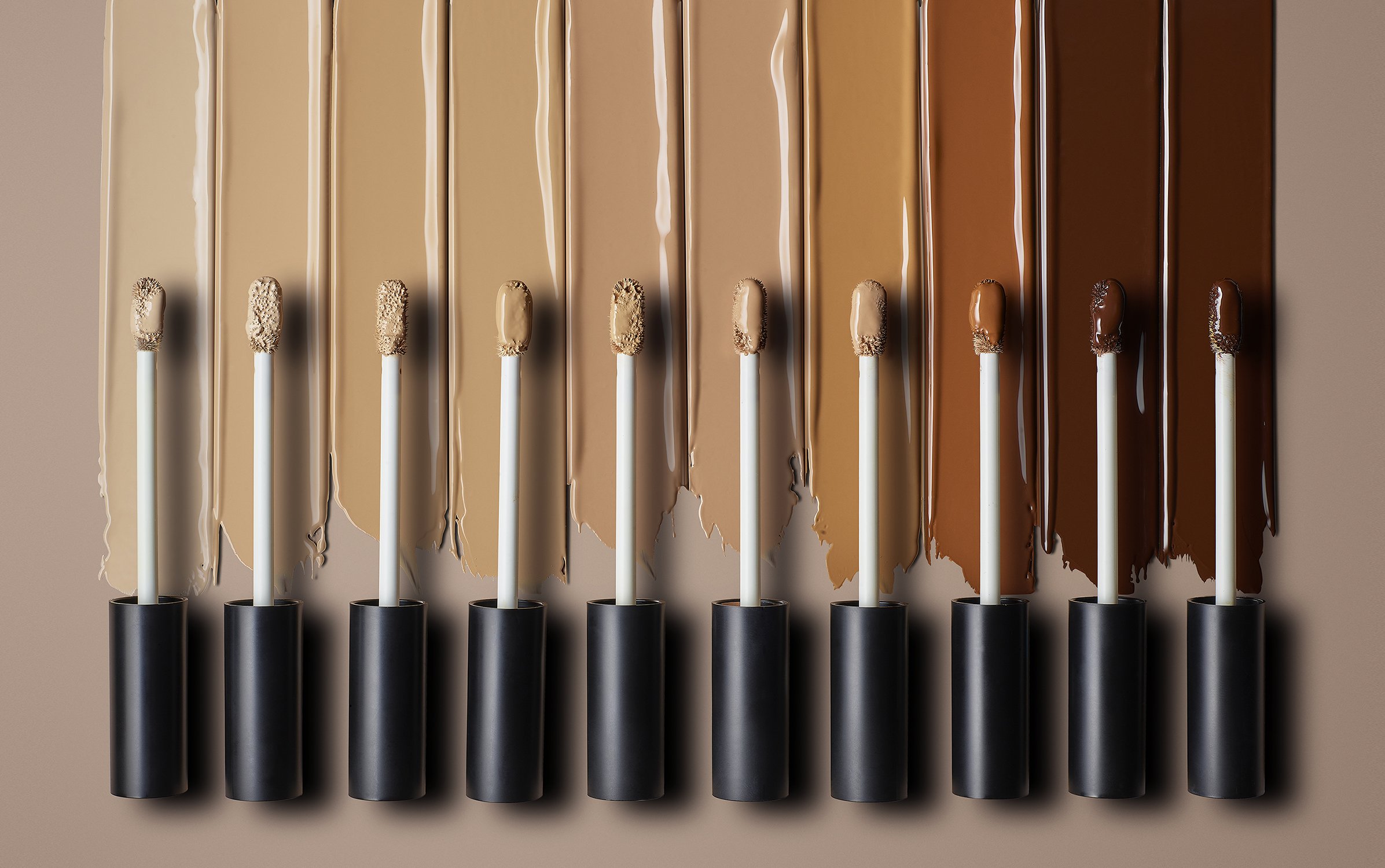 Close-up of various shades of liquid foundation swatches with applicators, ranging from light beige to dark brown on a neutral background.
