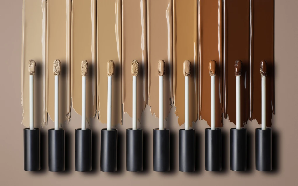 Close-up of various shades of liquid foundation swatches with applicators, ranging from light beige to dark brown on a neutral background.