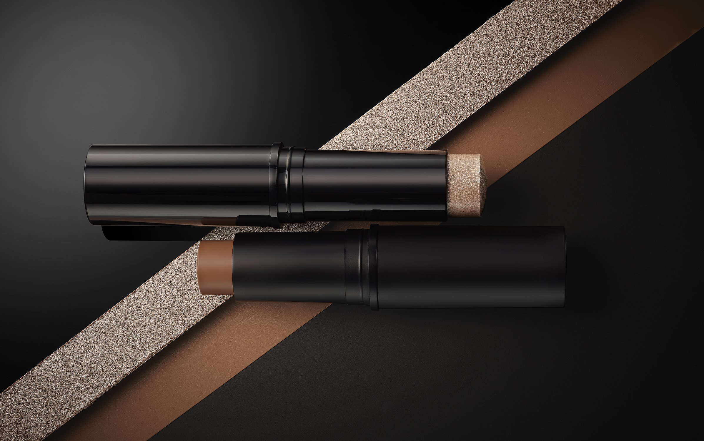 Two makeup contour sticks in black tubes are displayed against a dark background, with a textured ribbon adding depth to the composition.
