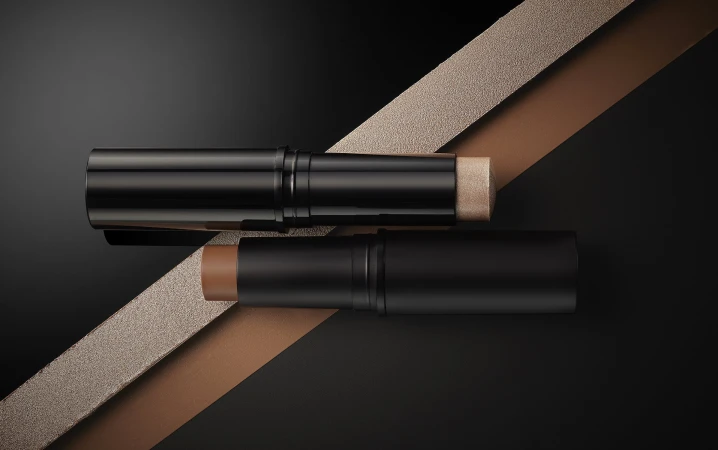 Two makeup contour sticks in black tubes are displayed against a dark background, with a textured ribbon adding depth to the composition.