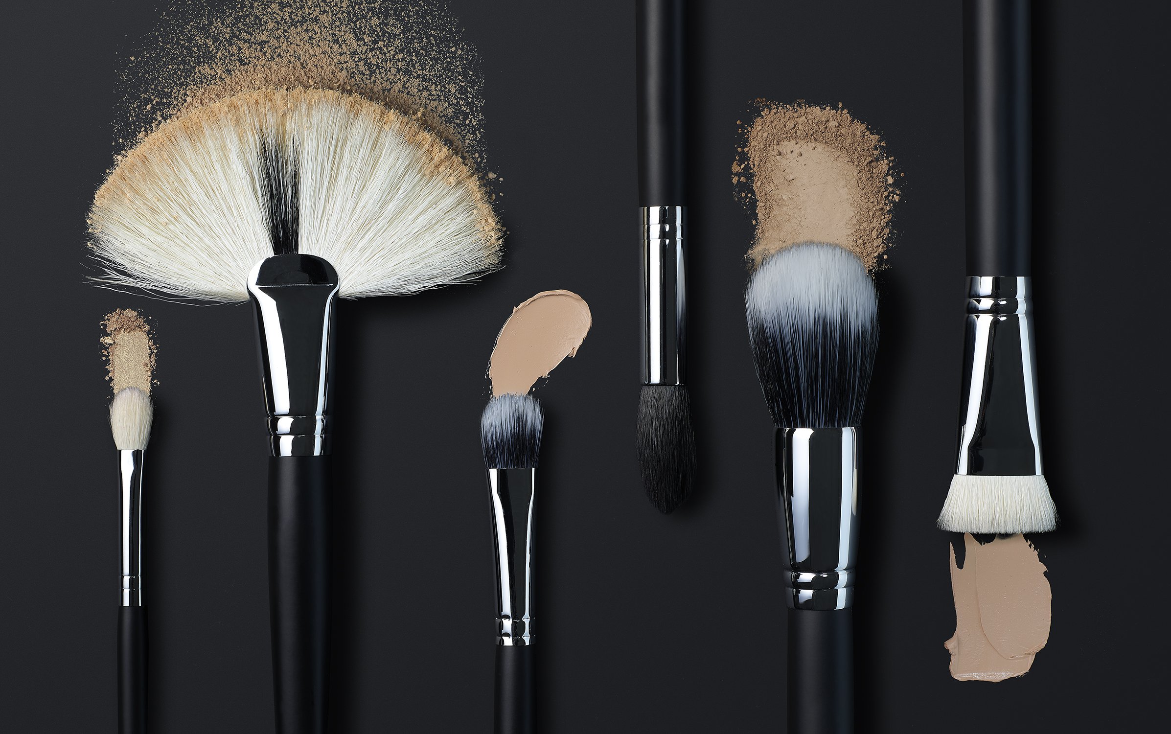 Arrangement of makeup brushes with dark handles and light bristles dusted with foundation powder against a dark background.