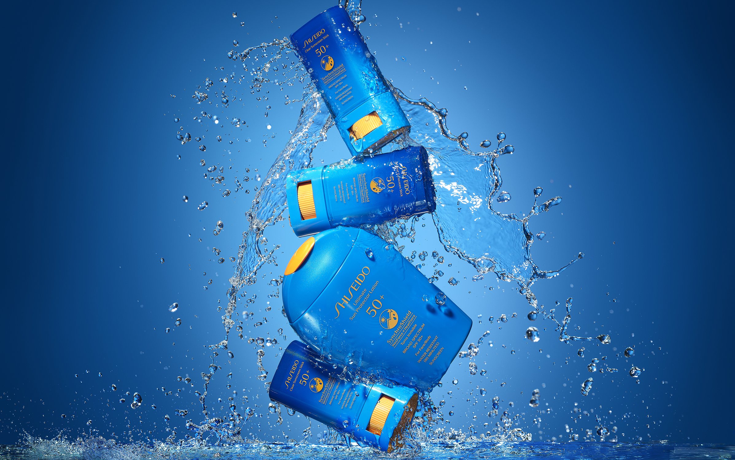 Four bottles of Shiseido SPF 50 sunscreen in mid-air, surrounded by water droplets, against a gradient blue background.