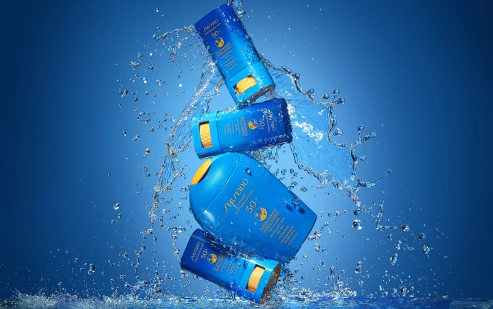 Four bottles of Shiseido SPF 50 sunscreen in mid-air, surrounded by water droplets, against a gradient blue background.